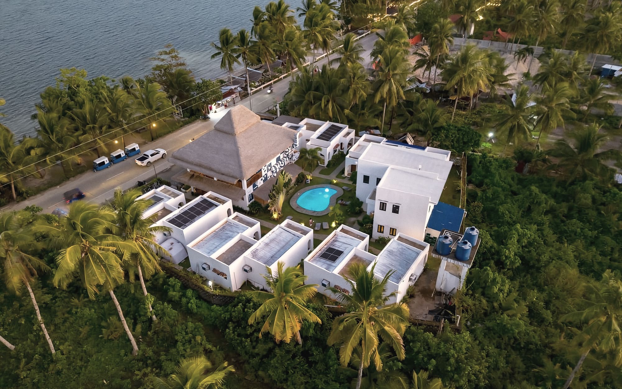 Aerial view of the hostel's white buildings, thatched main house and small pool among coconut palms, next to a coastal road and the sea.