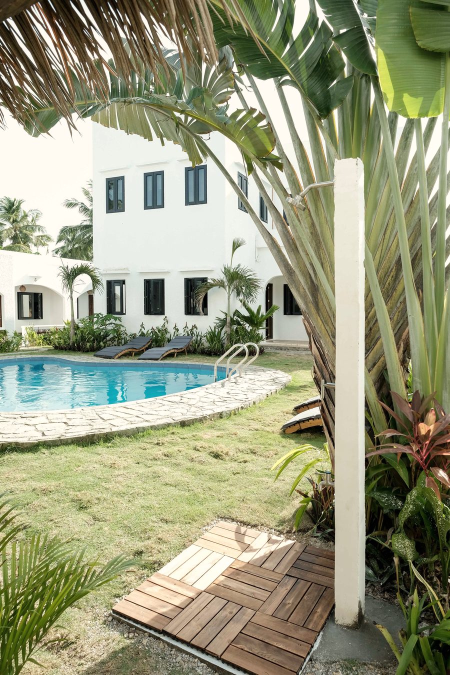 Outdoor shower on a wooden deck beside a traveller's palm, looking across the lawn to the courtyard pool, sun loungers and white buildings.