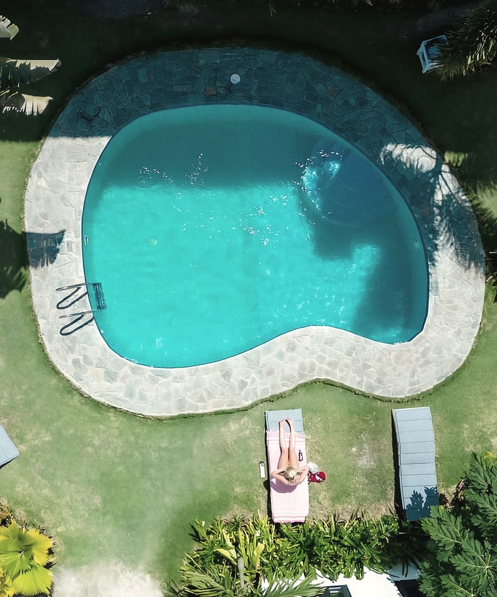 Top-down view of a kidney-shaped pool with stone edging on a lawn, with a person lying on a lounger beside it.