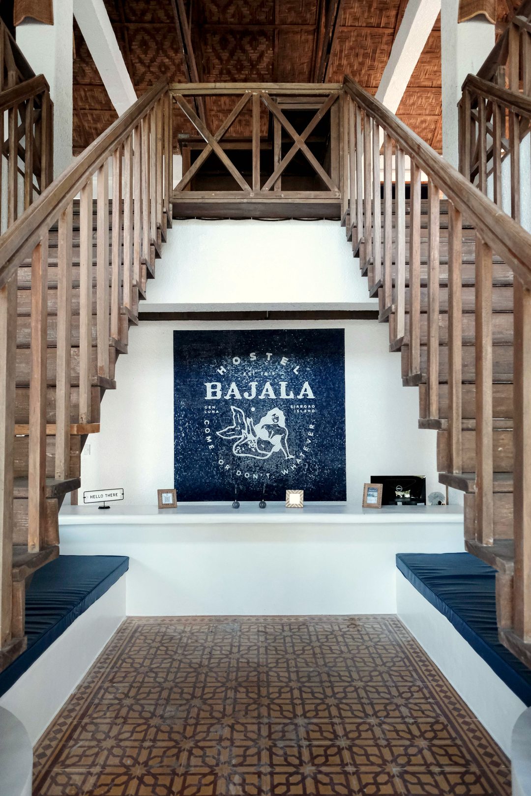 Reception counter under twin wooden staircases, with a navy Hostel Bajala banner on the white wall, navy cushioned benches either side and a patterned tile floor.