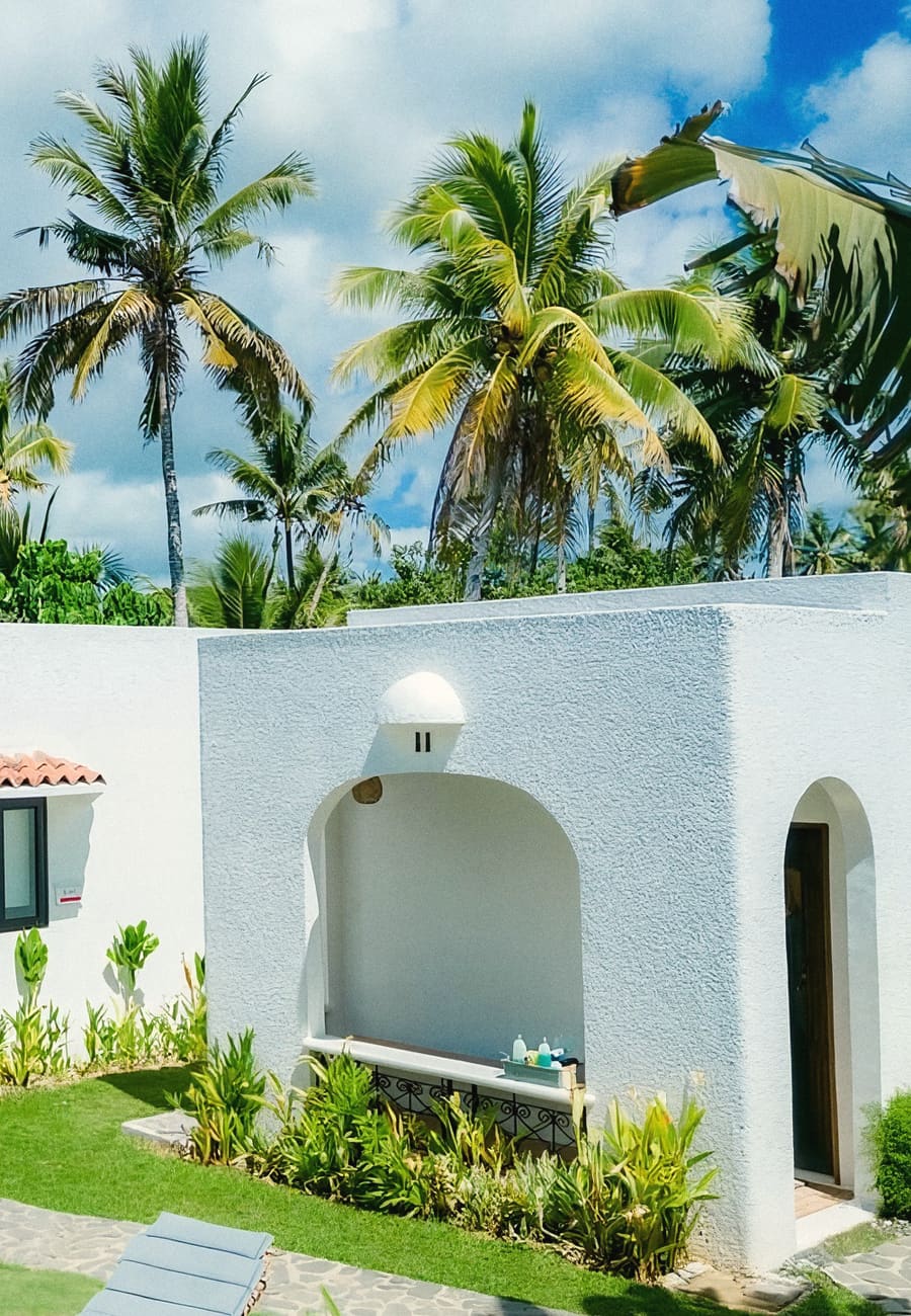 White arched cottages with a tiled roof and garden plants, with coconut palms and a blue sky above.
