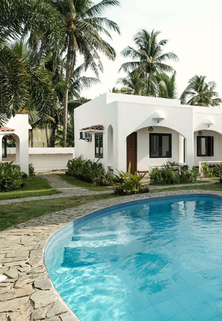 Curved pool with stone edging set in a lawn, with white cottages, sun loungers and coconut palms behind.