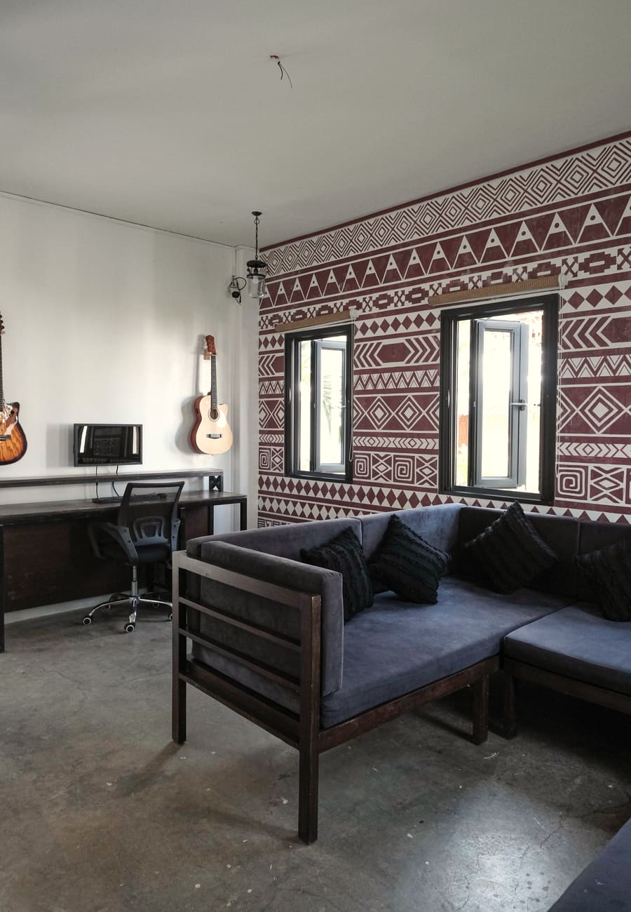 Lounge with a dark sofa, a desk with a computer monitor and office chair, guitars on the wall and a red-and-white patterned feature wall.