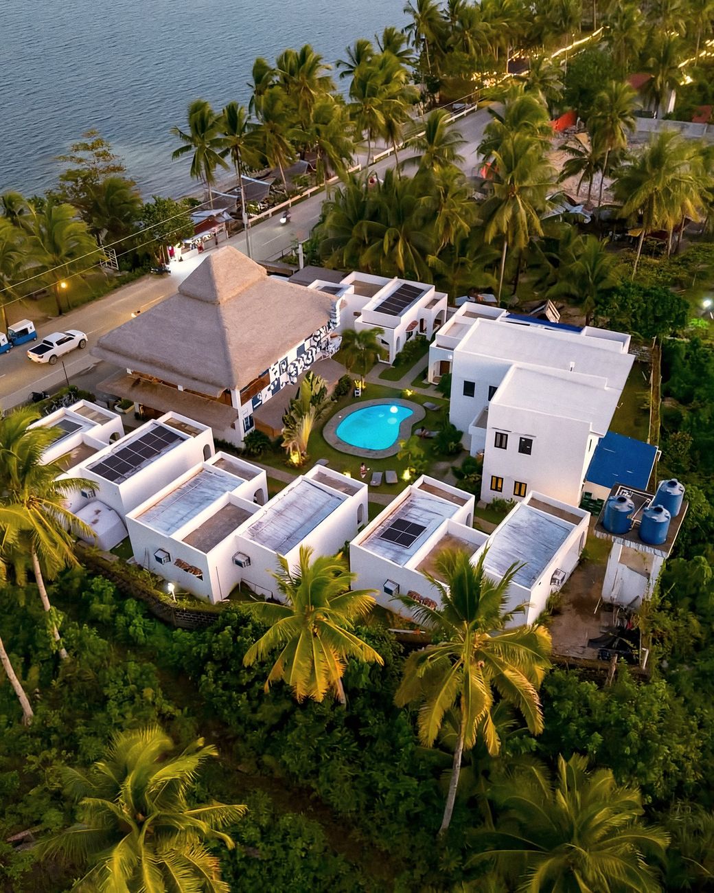 Aerial view at dusk of Bajala's white buildings, thatched main house and small blue pool among coconut palms, beside a coastal road and the sea.