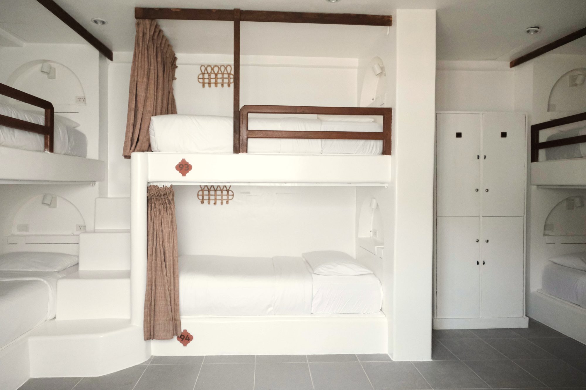Six-bed dorm with white built-in bunks, brown curtains, wooden rails and white lockers on a grey tiled floor.