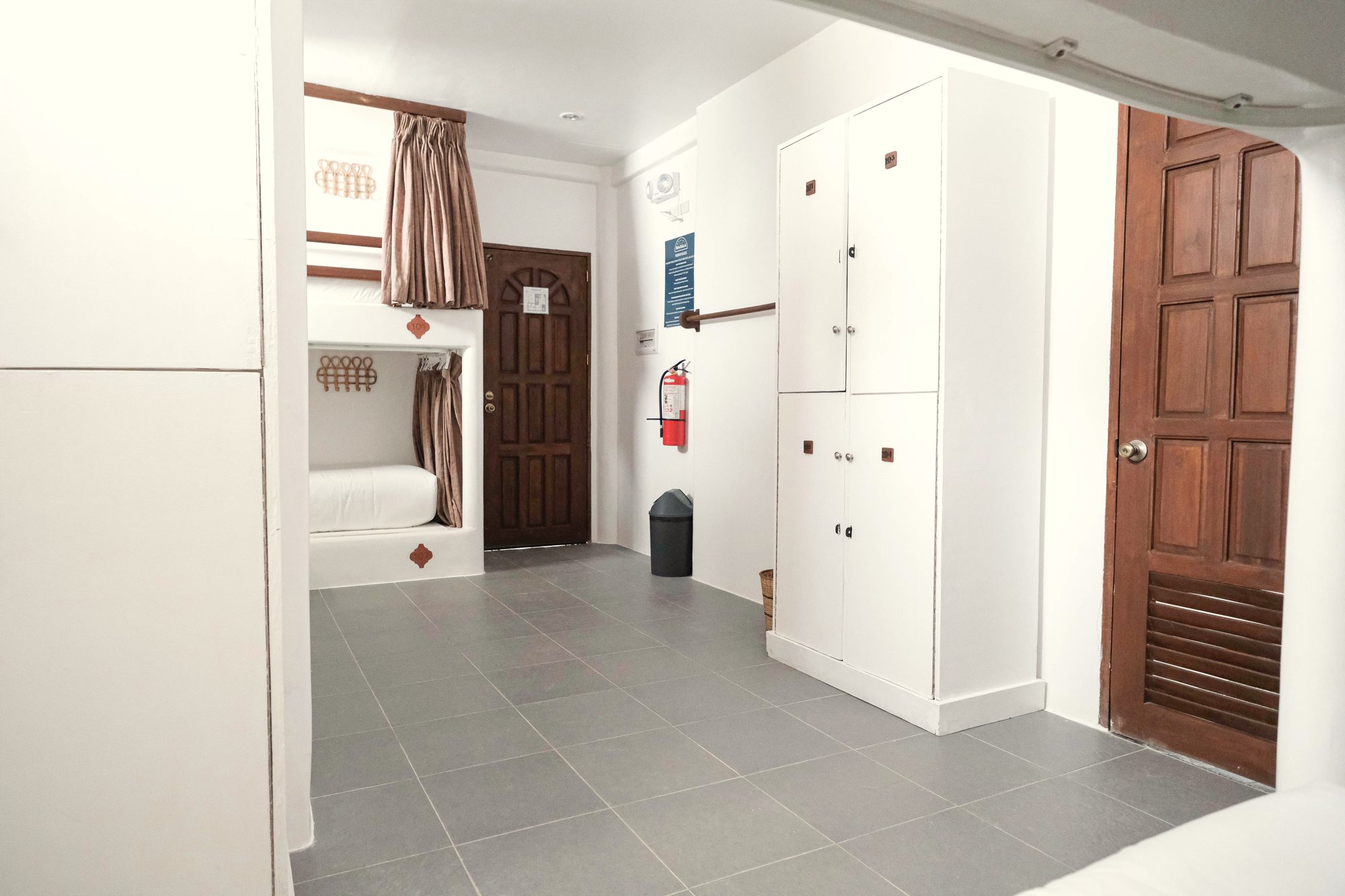 Mixed dorm seen from the doorway: bunks with brown curtains, tall white lockers, a carved wooden door and a grey tiled floor.