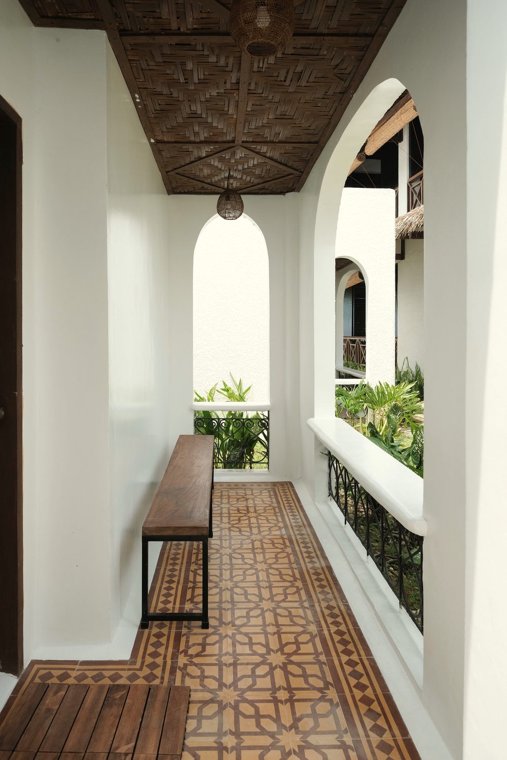 Arched veranda outside the rooms with a wooden bench, patterned tile floor, carved wooden ceiling and a pendant lamp.