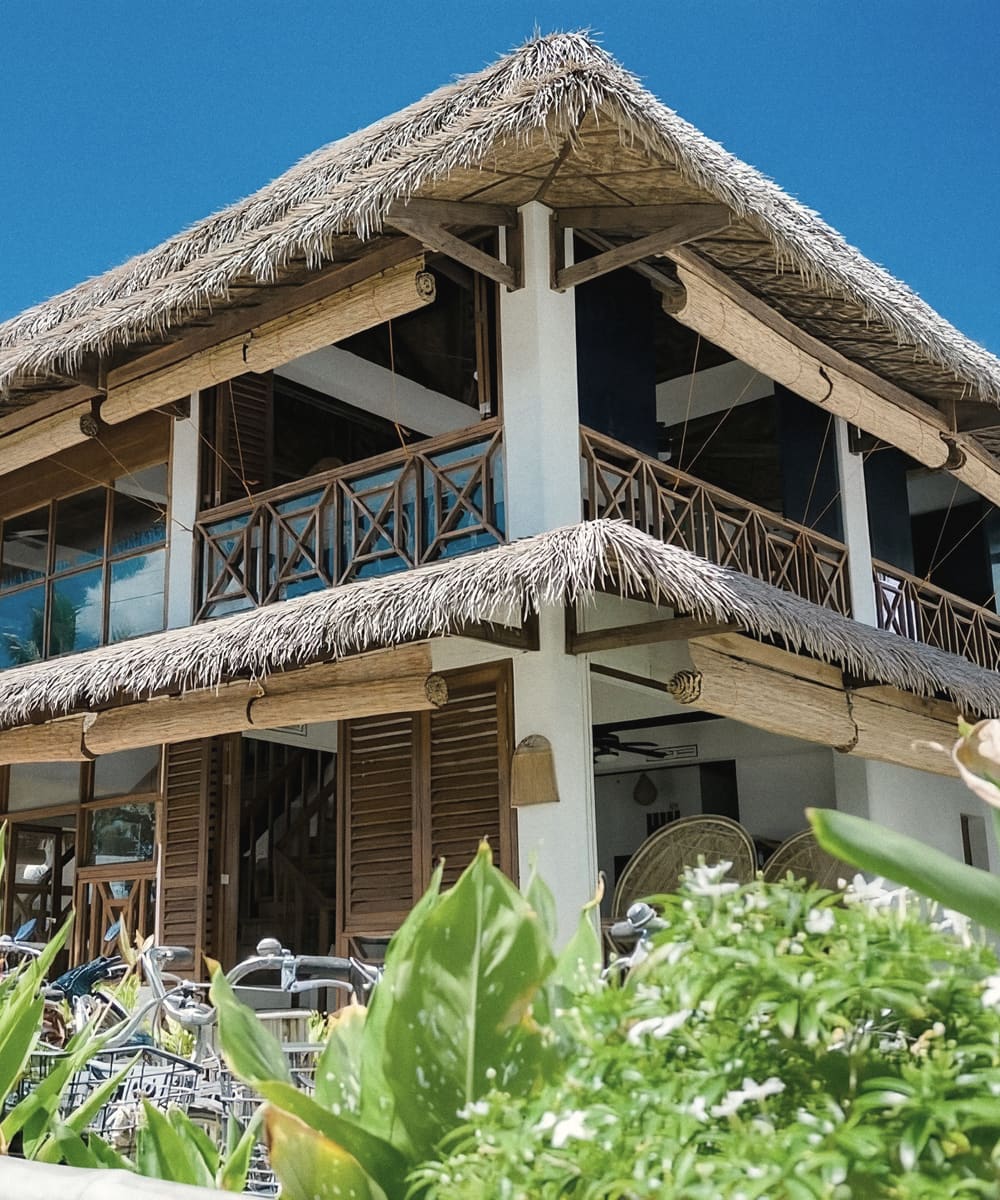Two-storey thatched-roof building with wooden balconies and shutters, behind flowering garden plants under a blue sky.