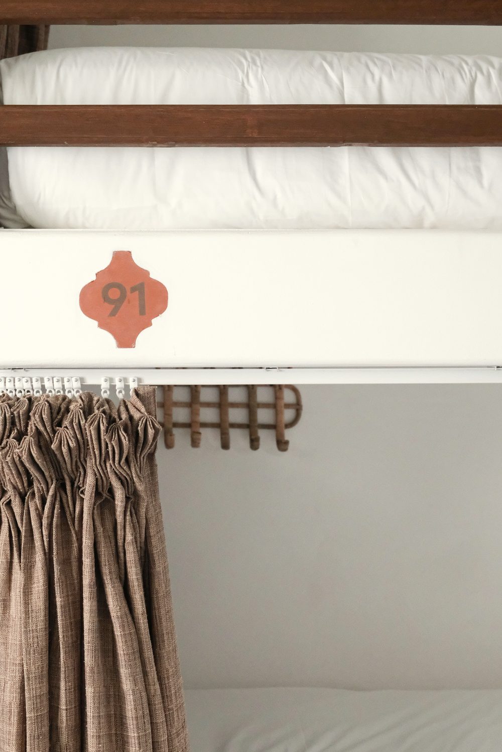 Upper and lower bunk with a wooden rail, a numbered terracotta tile, woven hooks and a brown privacy curtain.