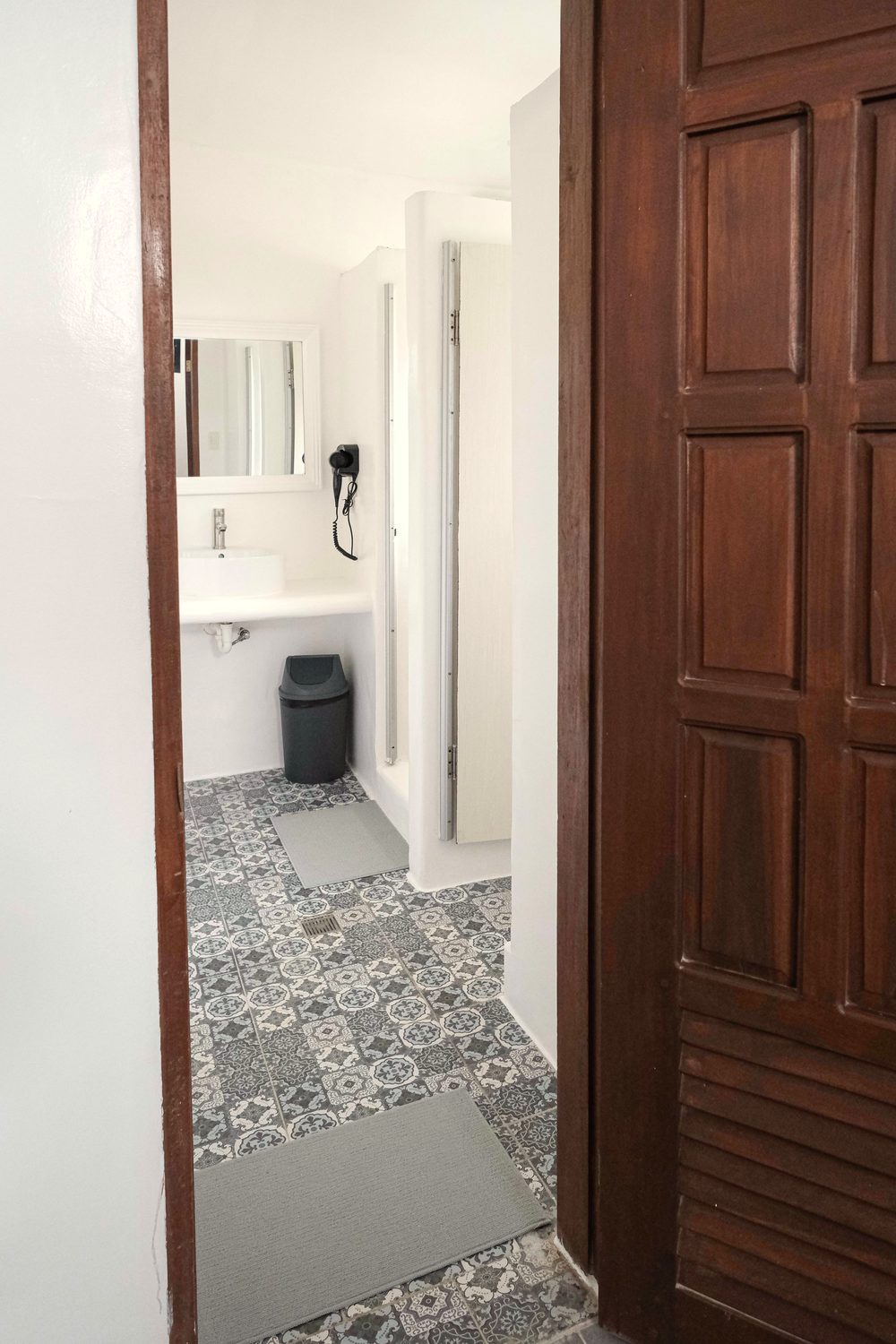 Ensuite bathroom seen through an open wooden door, with a basin, mirror, hair dryer and patterned floor tiles.