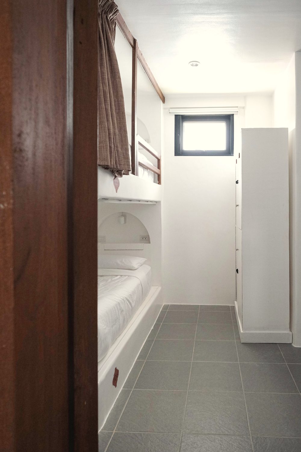 Narrow view past a dark wooden door into a dorm with white built-in bunks, brown curtains and tall white lockers.
