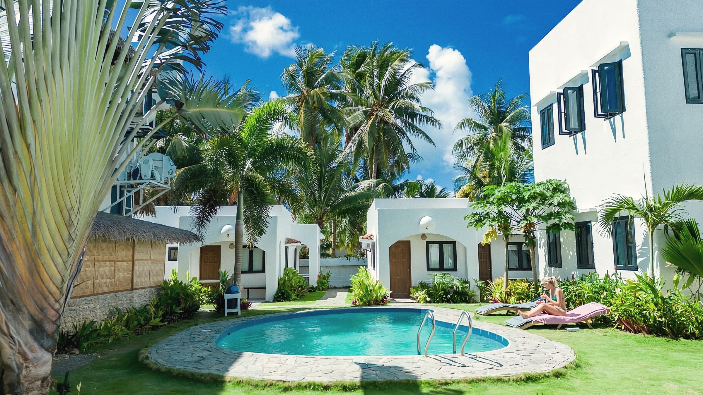 Oval swimming pool in a grassy courtyard surrounded by white cottages, a white multi-storey building and coconut palms under a blue sky; a guest lies on a sun lounger by the pool.