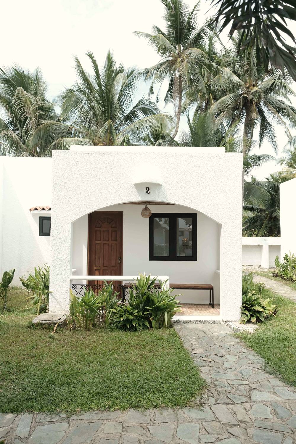 White arched cottage porch with a wooden bench, a carved door and a black-framed window, reached by a stone path across the lawn.