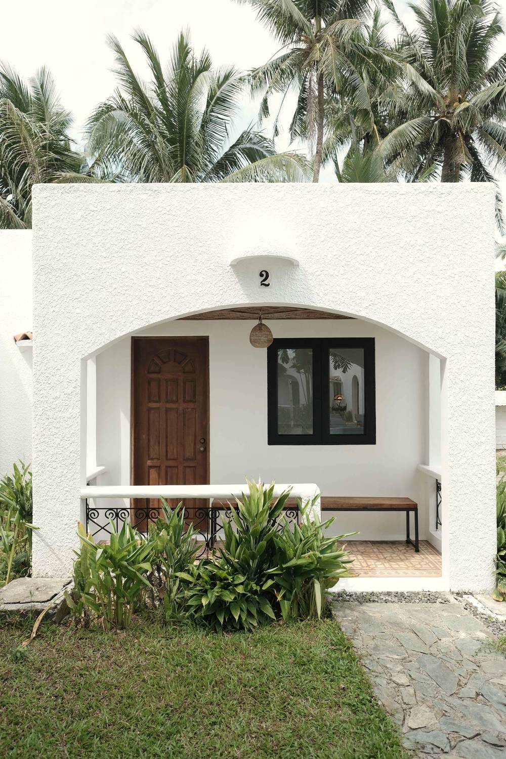 White arched cottage with a wooden door and a black-framed window, set behind a garden.