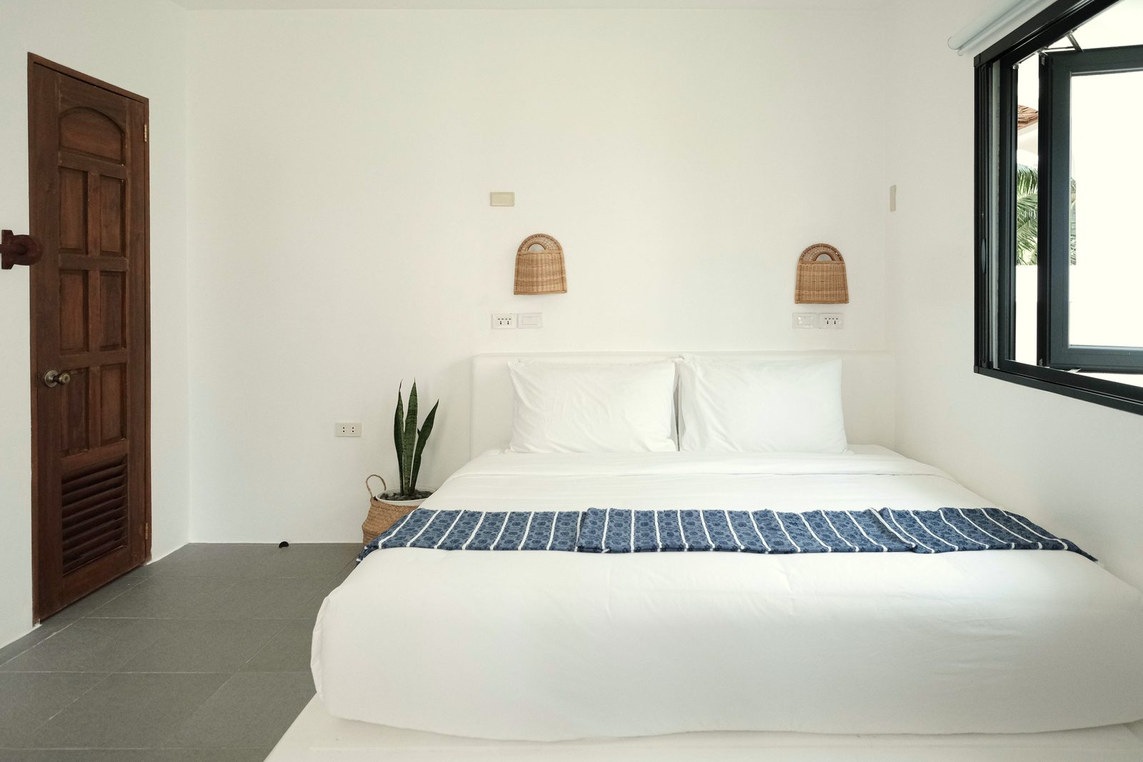 King bed with white linen and a blue striped runner between two woven wall lamps, with a carved wooden door on the left and a black-framed window on the right.