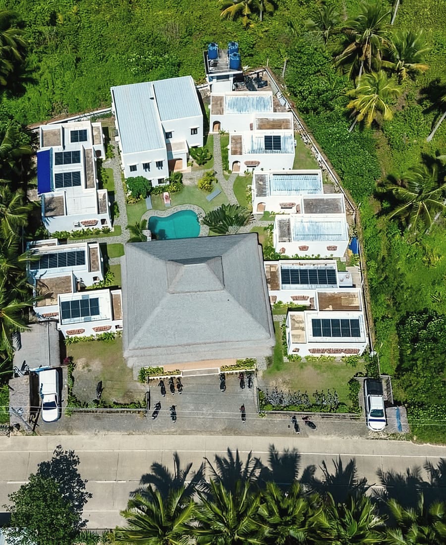Top-down aerial view of the hostel's white buildings with solar panels, a small pool and a large grey roof among palm trees beside a road.