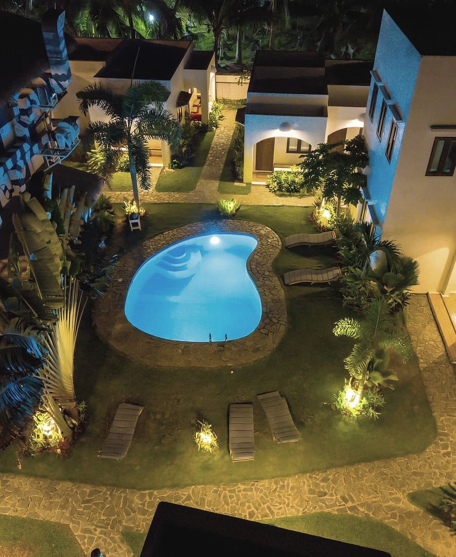 Courtyard pool lit at night seen from above, surrounded by lawn, loungers, palms and white buildings.
