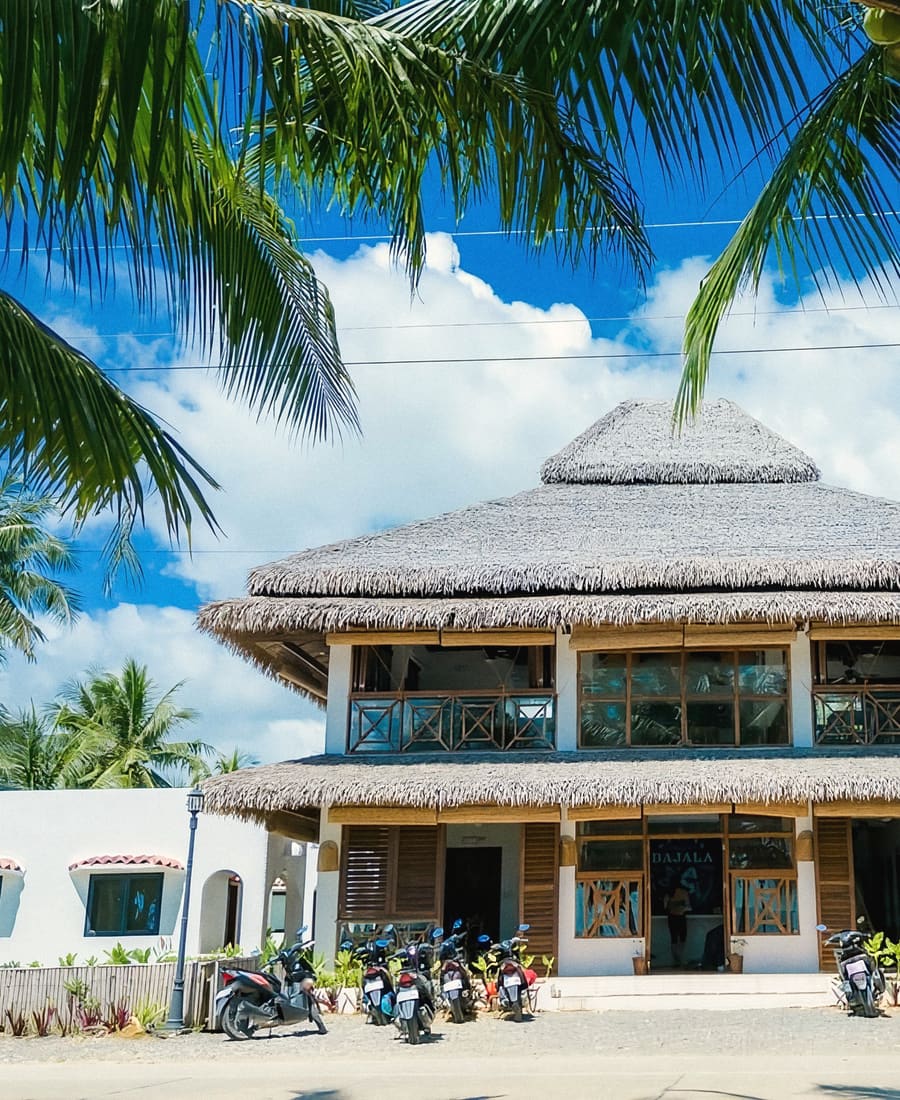 Thatched-roof main building with wooden balconies, framed by palm fronds, with motorbikes parked in front.