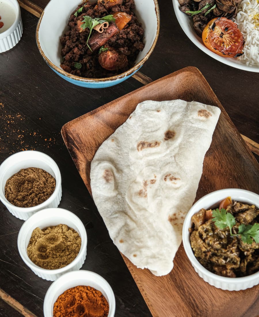 Overhead view of flatbread on a wooden board, bowls of curry and three small bowls of spices on a dark table.