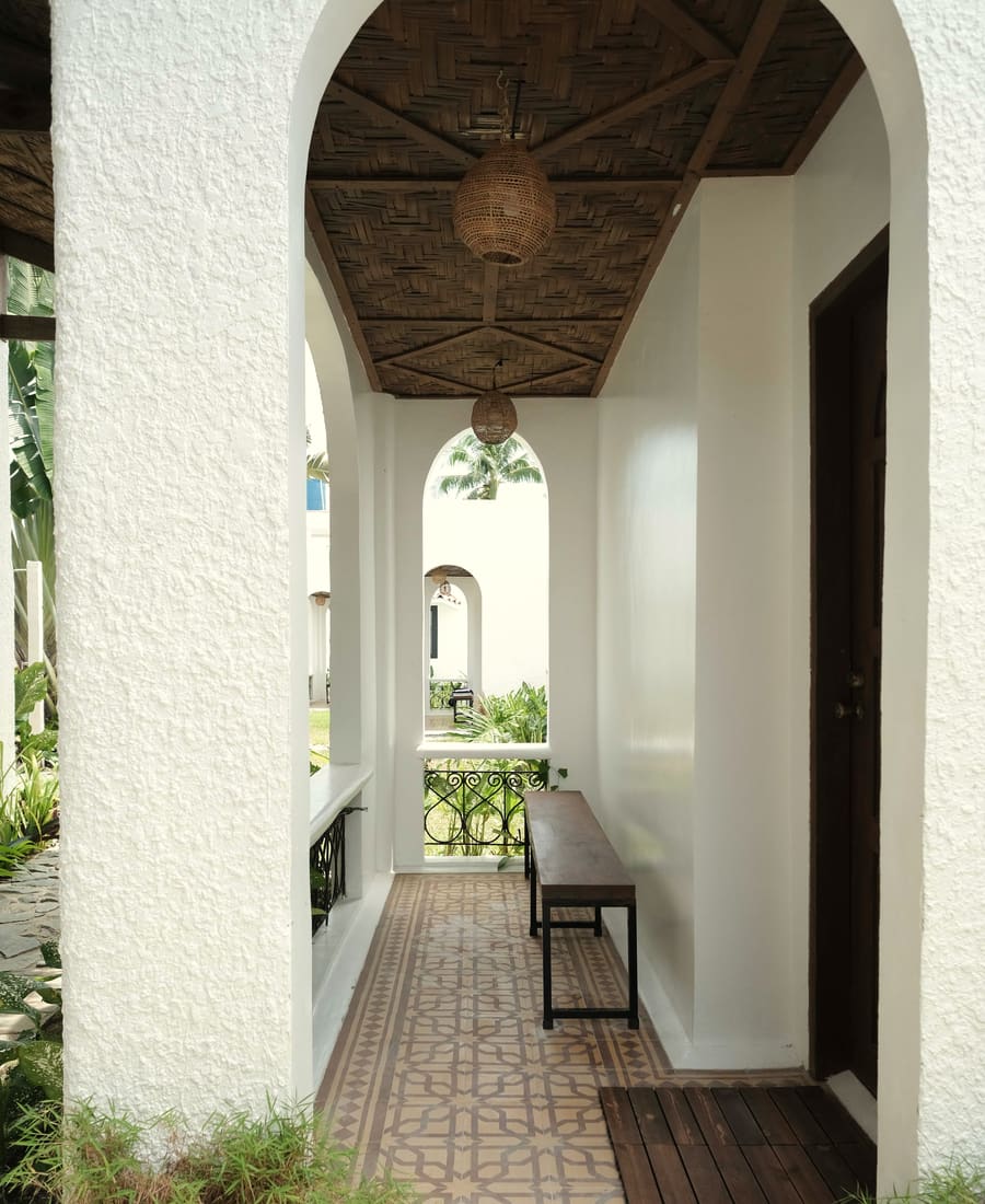 Arched veranda with a patterned tile floor, a wooden bench, a woven ceiling and a pendant lamp beside a room door.