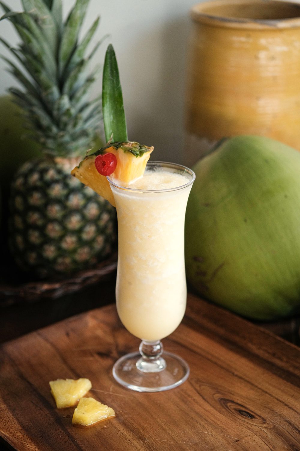 Creamy blended cocktail in a tall glass garnished with pineapple and a cherry, on a wooden board beside a whole pineapple and a green coconut.