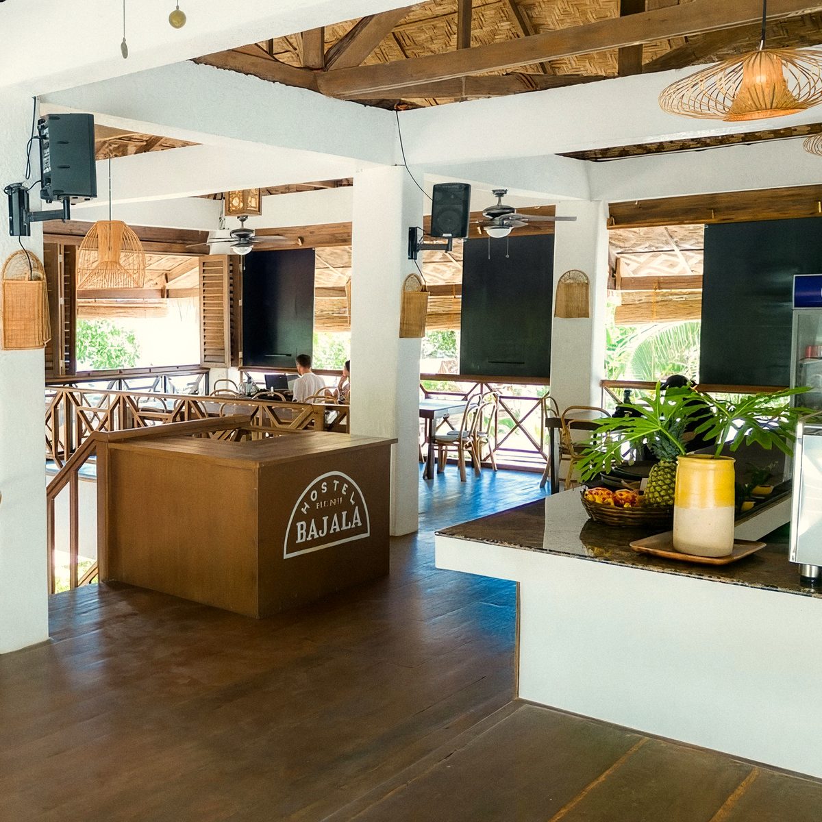 Little Rotti's open-sided dining room with a wooden counter marked Hostel Bajala, a dark wooden floor, woven pendant lamps and tables beyond.