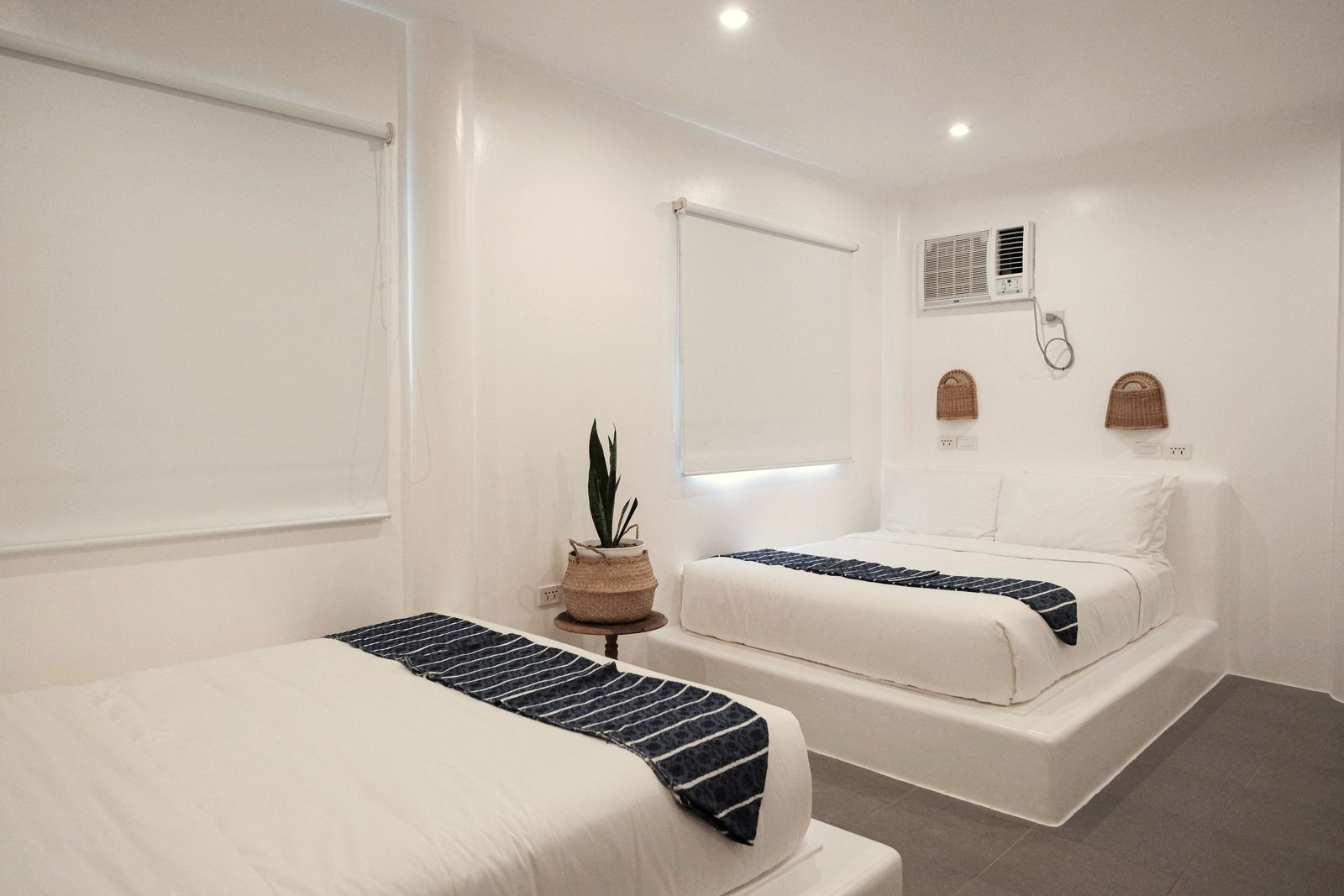 Private Quad with a double bed under a window blind, a second bed in the foreground, a snake plant and a wall-mounted air-conditioner.
