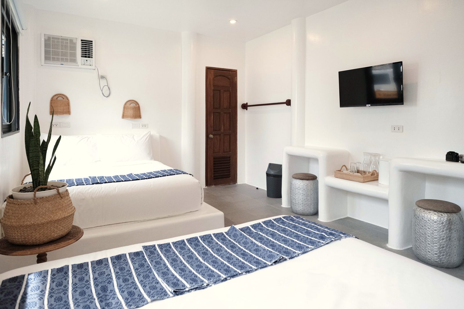 Private Quad with two double beds with blue striped runners, a snake plant in a basket, woven wall lamps and a carved wooden door.