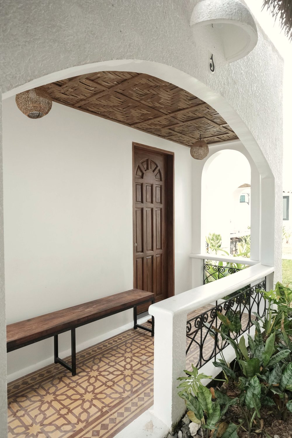 Arched porch with a carved wooden door, a wooden bench, a patterned tile floor and a woven ceiling.