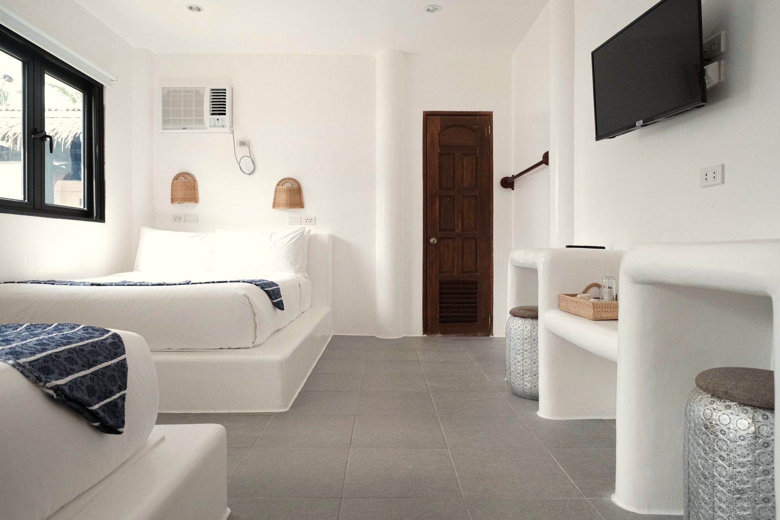 Private Quad seen from the window side: two double beds, a white built-in desk with stools, a wall-mounted TV and a carved wooden door.