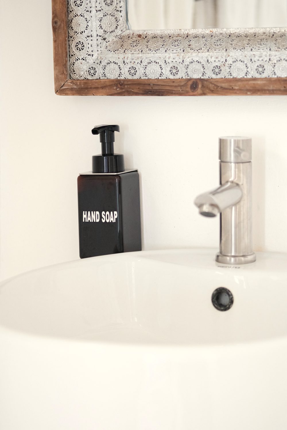 White basin with a steel tap and a black hand-soap dispenser below a mirror with a patterned tile frame.