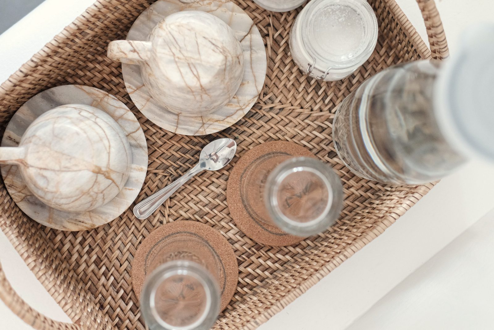 Woven rattan tray with marbled ceramic cups, glasses, coasters and a teaspoon on a white surface.