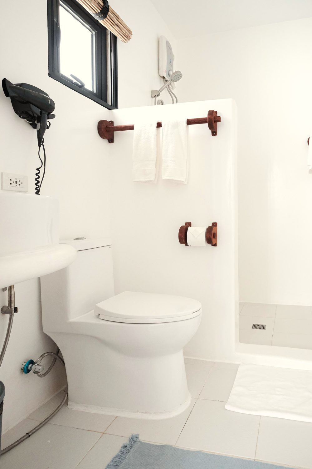 White ensuite bathroom with a basin, wall-mounted hair dryer, towels on a wooden rail, a toilet and a small black-framed window.