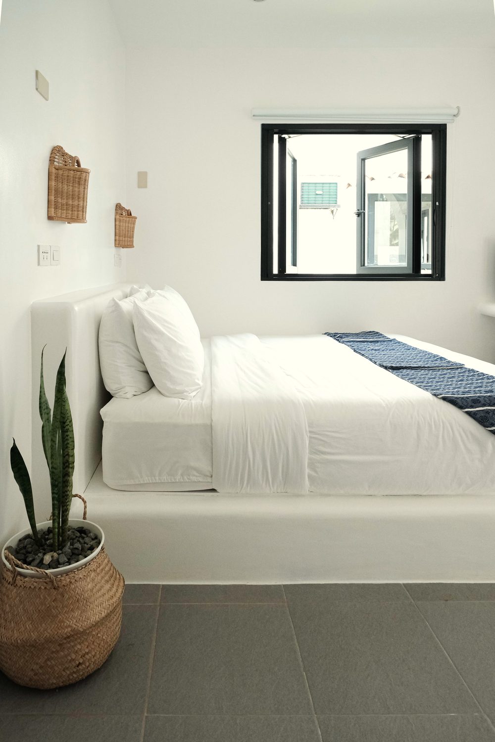 Queen bed with white linen and a blue striped runner beside a black-framed window, with a woven wall lamp and a snake plant in a basket.