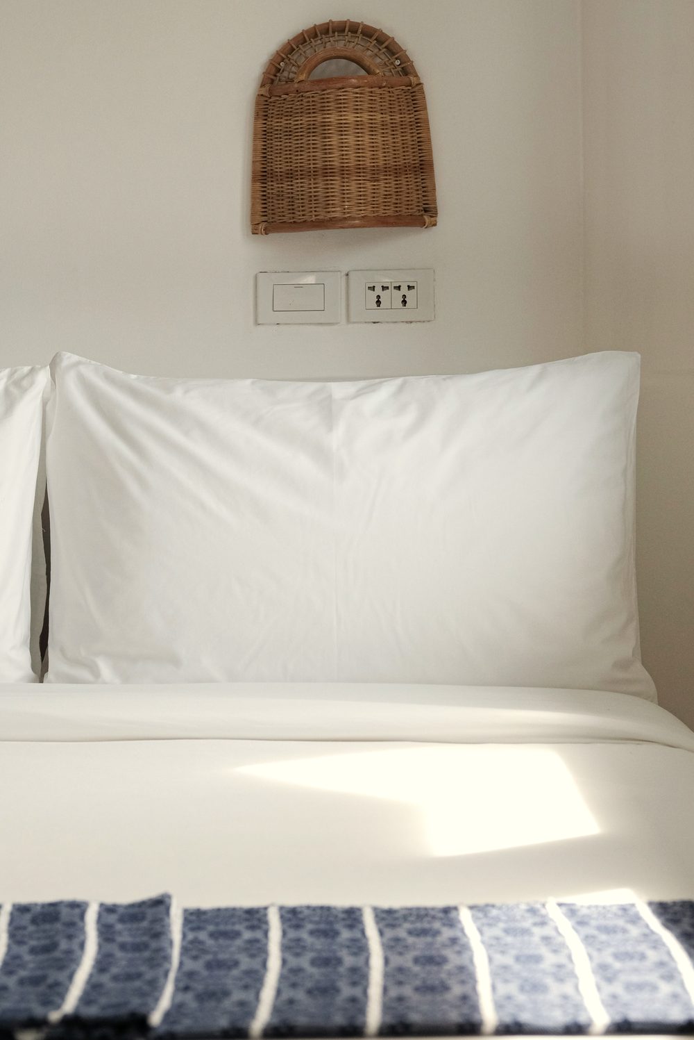 Close-up of white pillows and a blue patterned runner on the queen bed, under a woven wall lamp and two sockets.