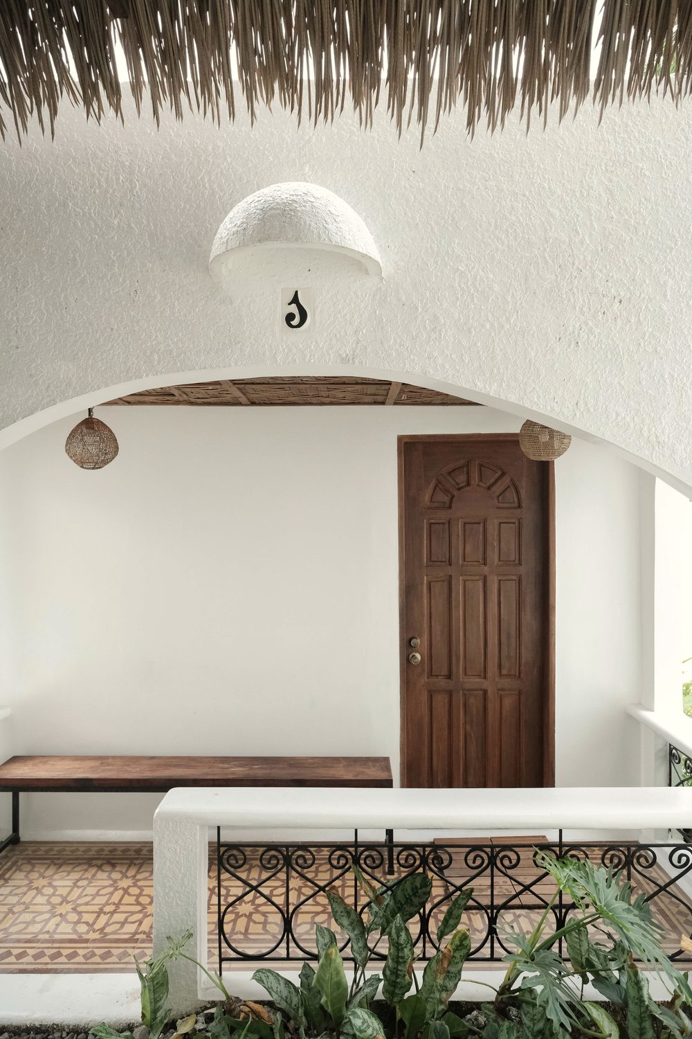 Arched porch outside a private room with a carved wooden door, a wooden bench, a wrought-iron railing and a woven pendant lamp.