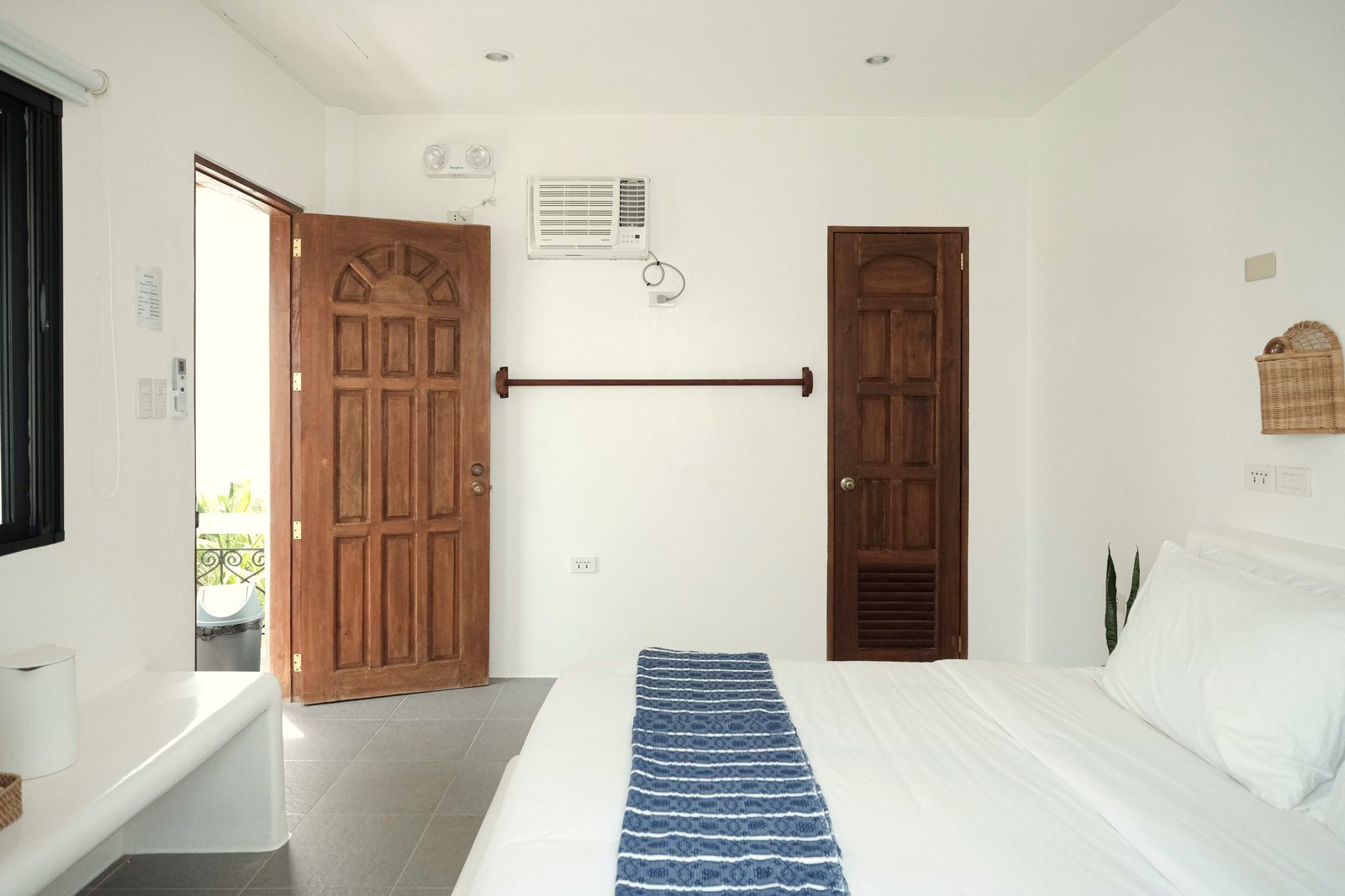 Private room with a carved wooden entrance door, a second wooden door, a wall-mounted air-conditioner and the end of a bed with a blue striped runner.