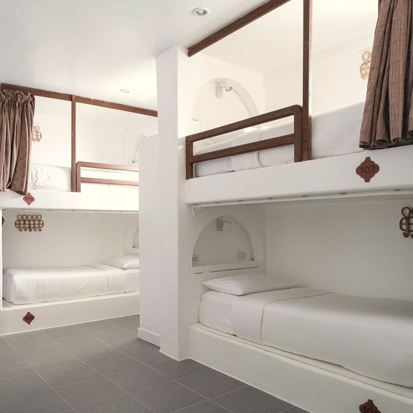 Dorm room with white bunk beds, wooden ladders and rails, brown curtains and a grey tiled floor.