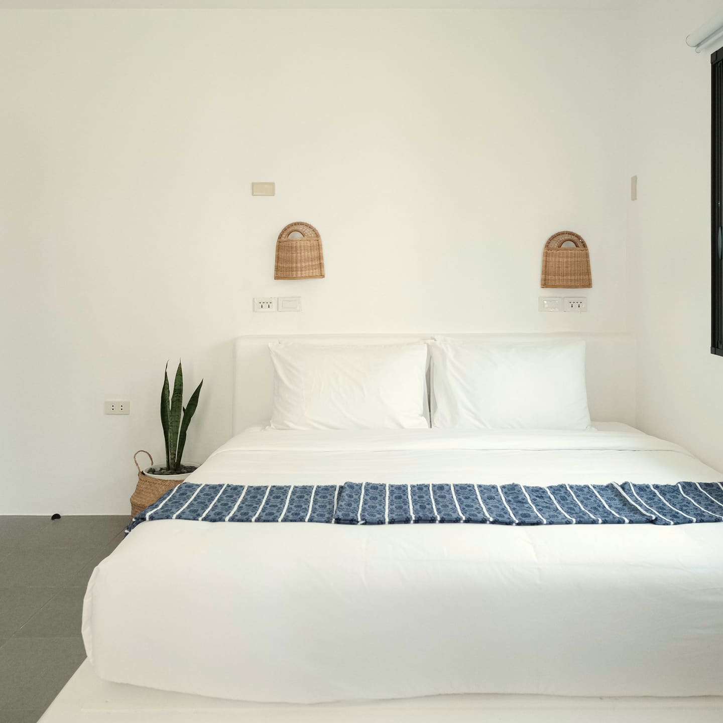 Double bed with white linen and a blue striped runner, woven wall lamps, a carved wooden door on the left and a black-framed window on the right.