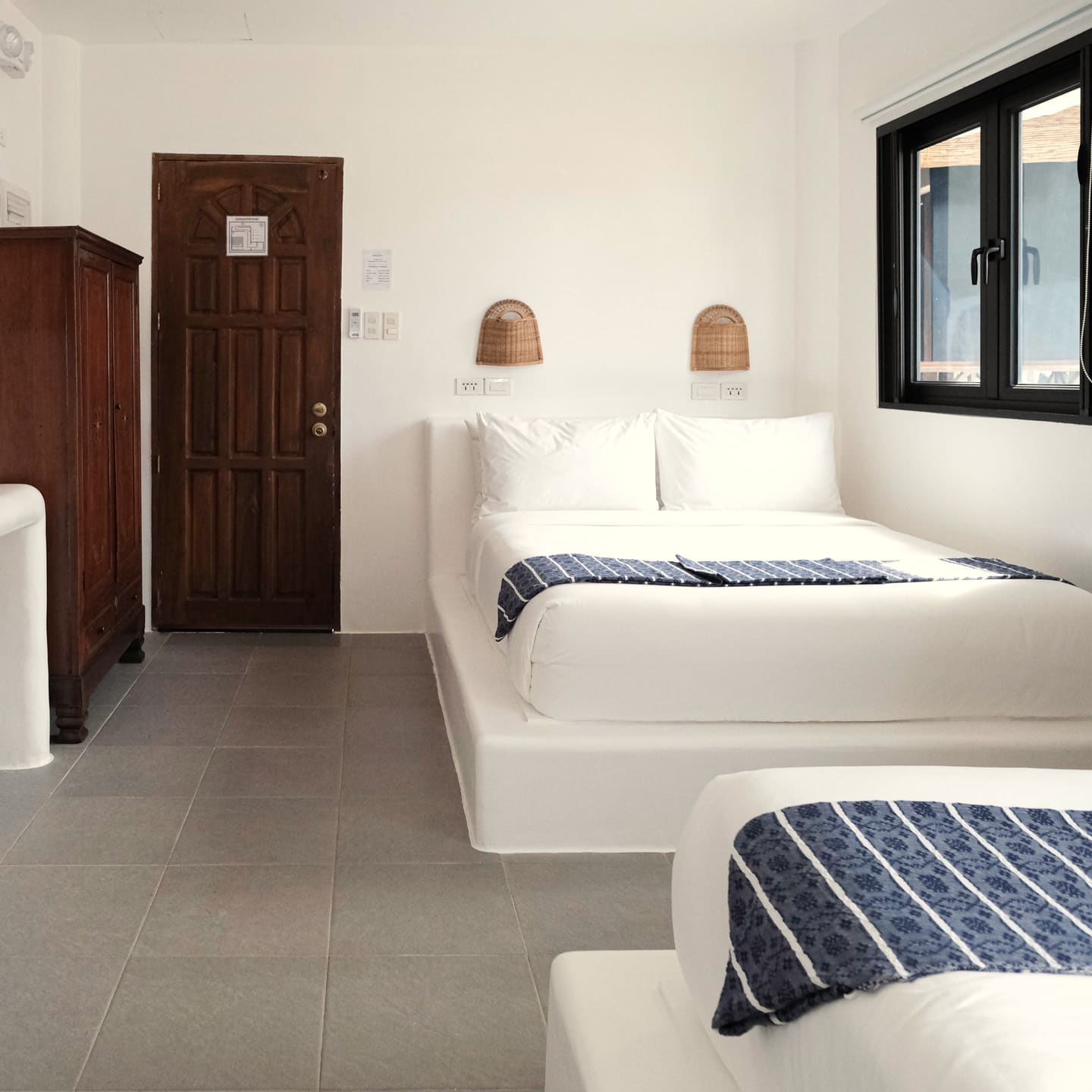 Room with two double beds, a dark wooden wardrobe, a carved door, woven wall lamps and a black-framed window over a grey tile floor.