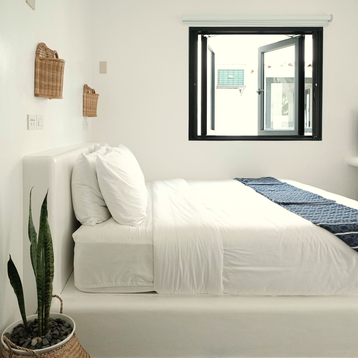 Double bed with white linen and a blue striped runner under a black-framed window, with woven wall lamps and a snake plant.