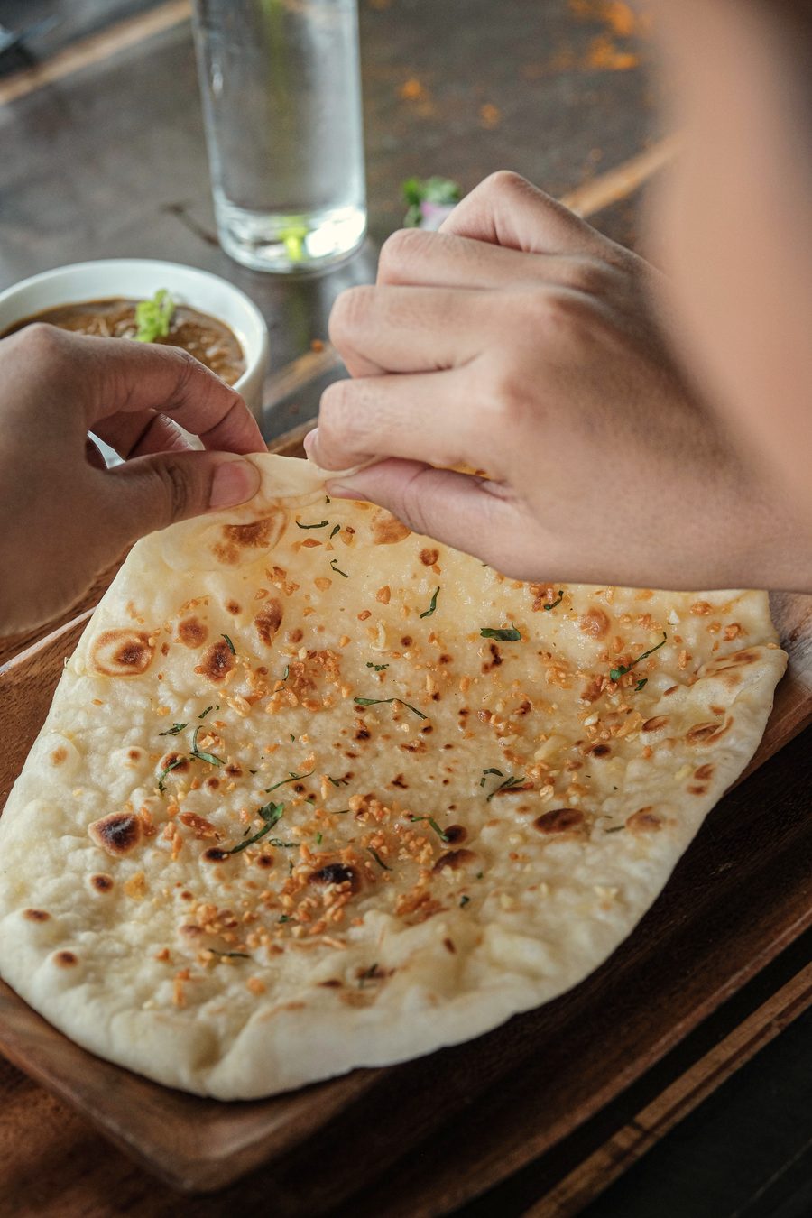 Hands tearing a garlic naan on a wooden board, with a glass of water behind.