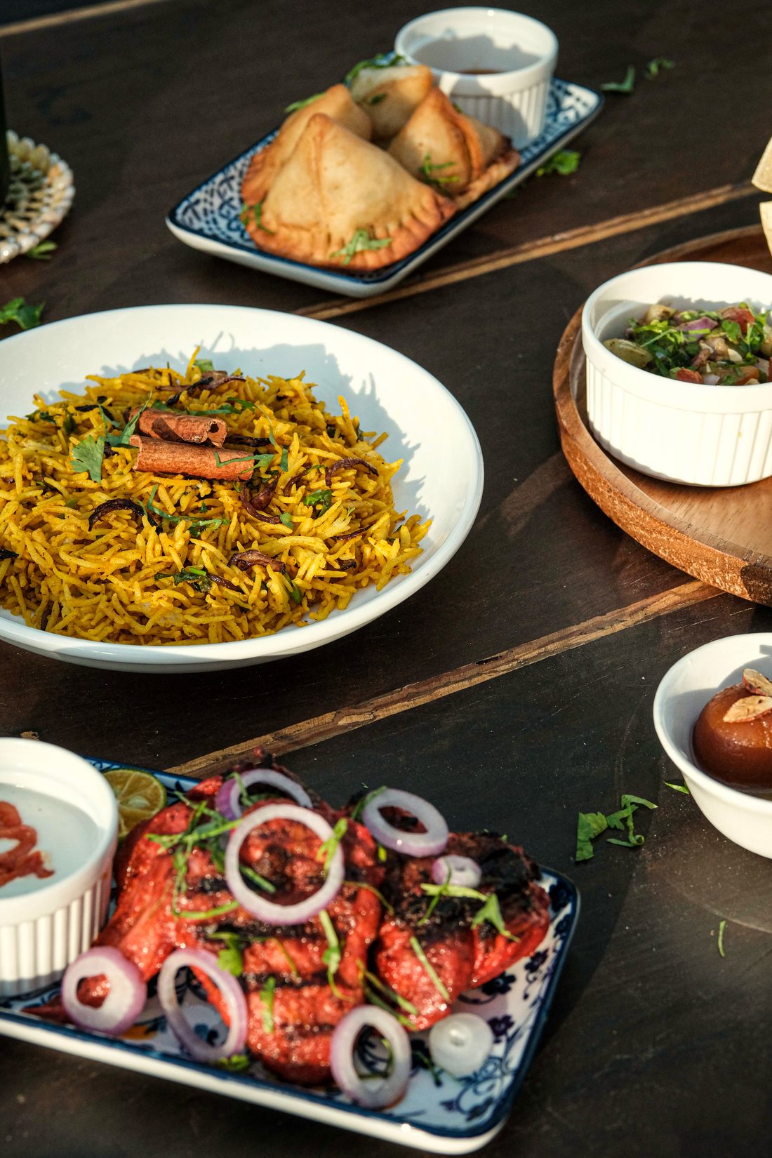 Dark wooden table seen from above with a platter of samosas, a bowl of yellow biryani, tandoori chicken with onion rings and lime, and small bowls of curry.