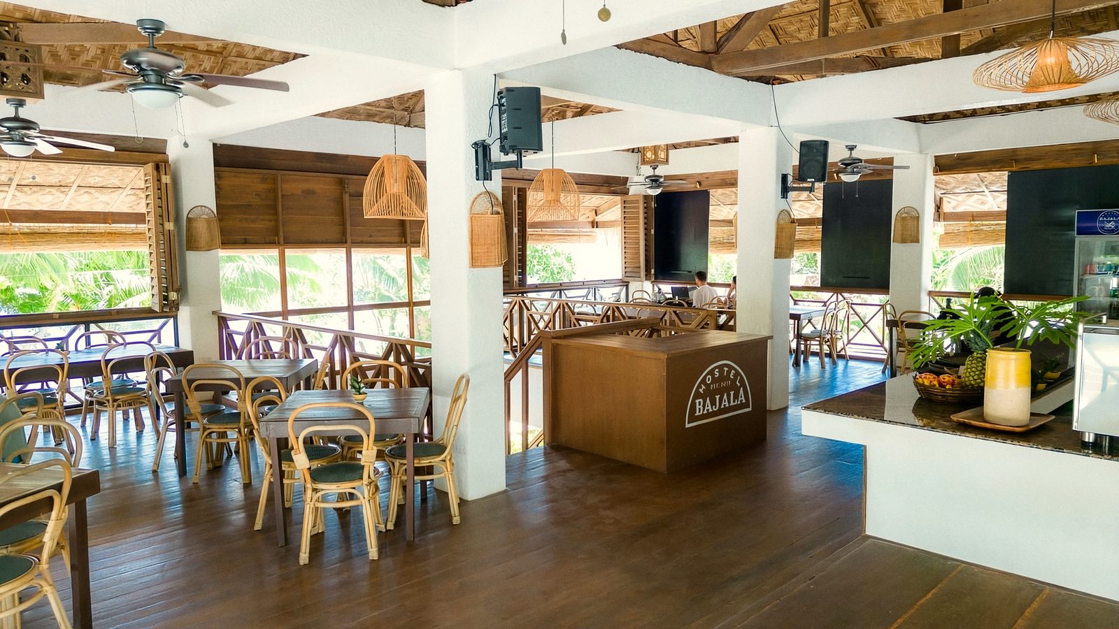 Open-sided restaurant under a woven ceiling, with wooden tables and rattan chairs, ceiling fans, potted plants and a white counter.