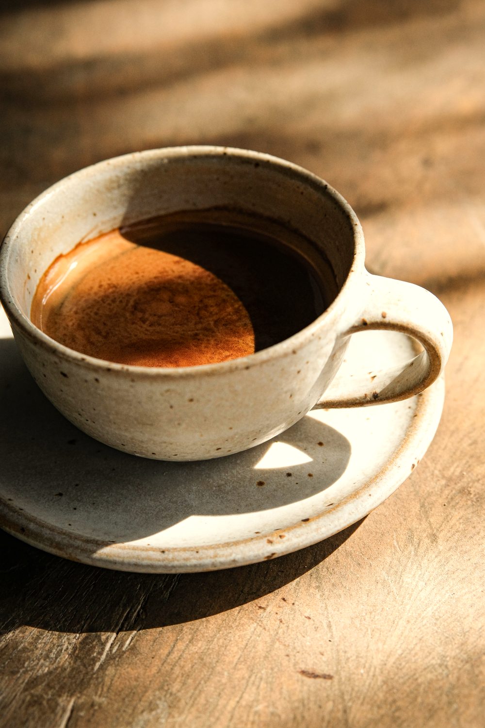 Black coffee in a speckled ceramic cup and saucer on a wooden table in warm morning light.