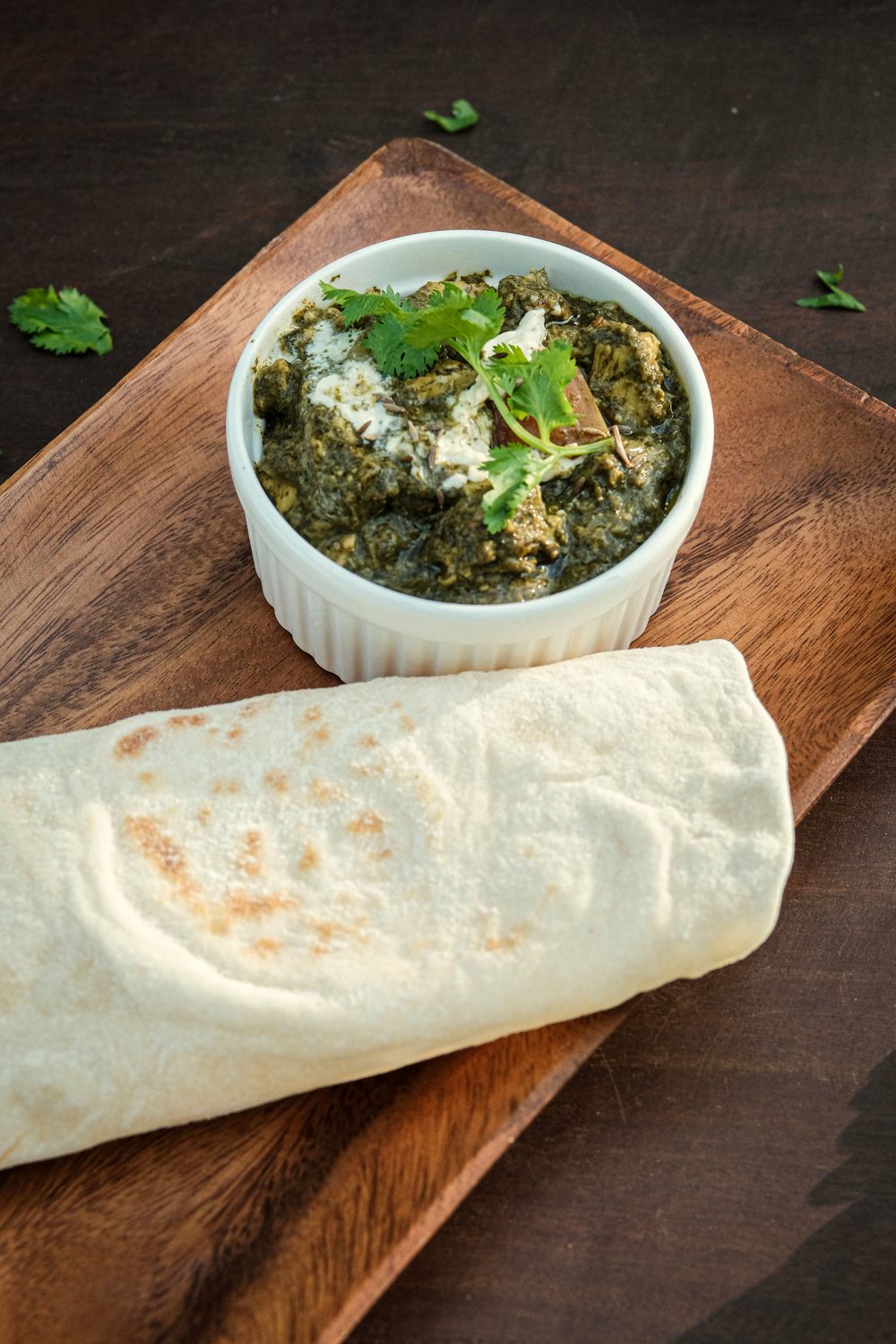 Bowl of green spinach curry topped with cream and coriander beside a rolled flatbread on a wooden board.