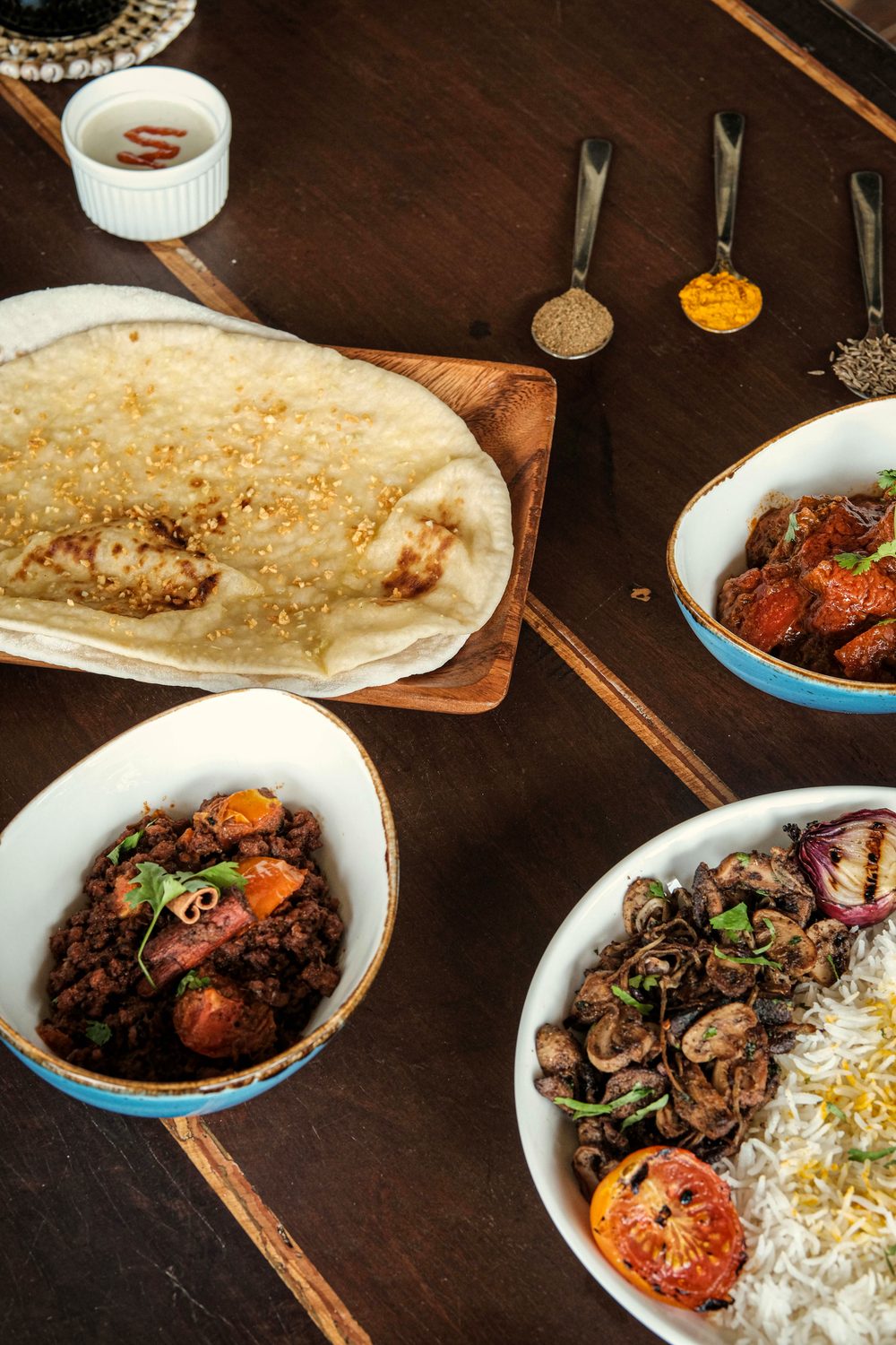 Overhead view of a curry spread on a dark table: naan on a wooden board, bowls of chicken curry, rice with fried onions and small spoons of spices.