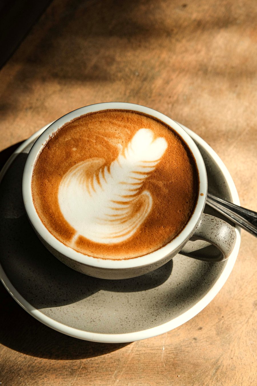 Cappuccino with latte art in a dark grey cup and saucer on a wooden table in morning sun and leaf shadows.
