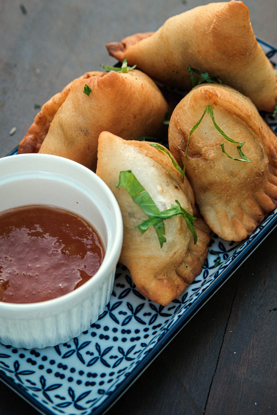 Golden samosas on a blue-and-white patterned plate with a small bowl of red dipping sauce.
