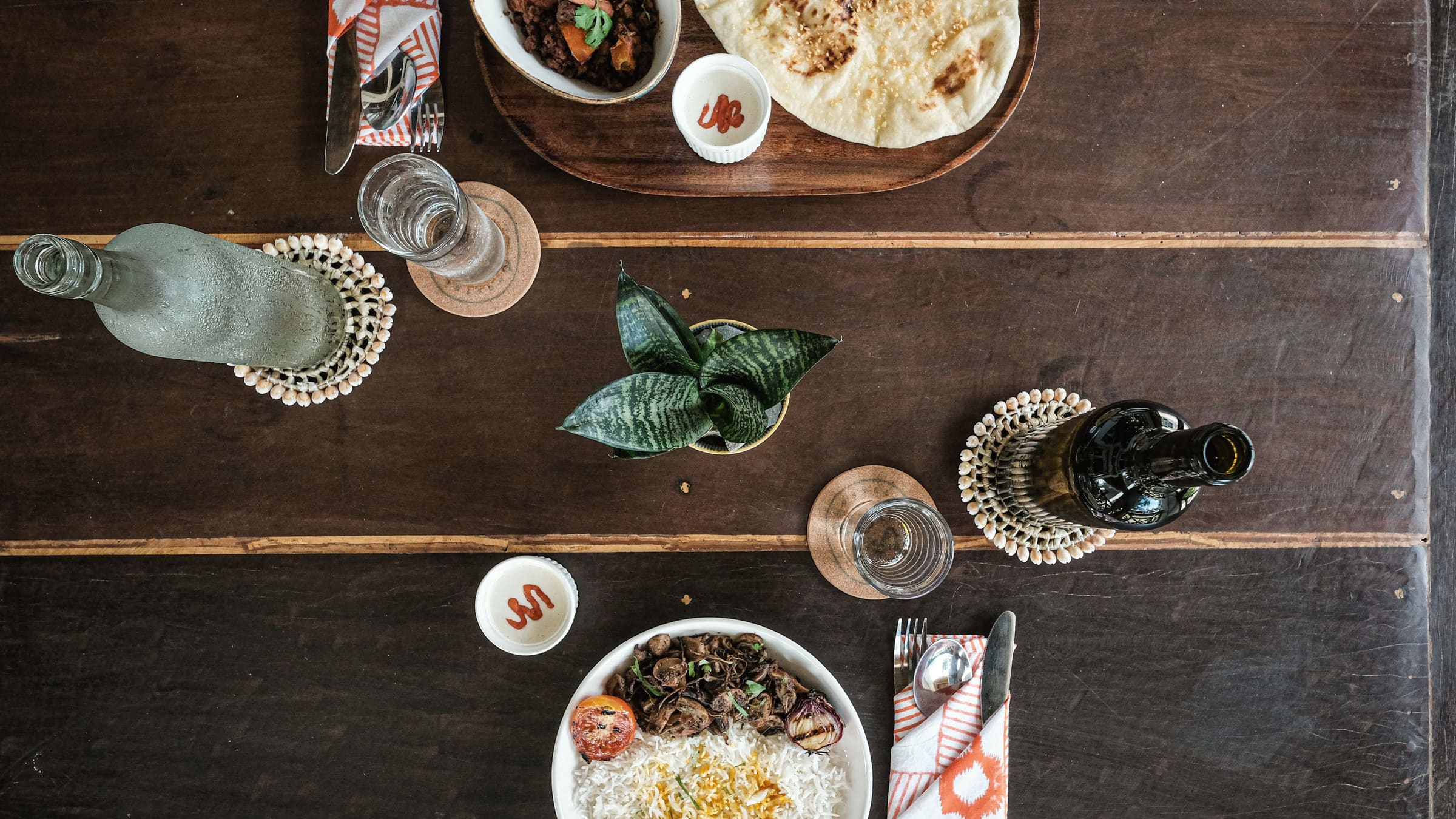 Dark wooden table seen from above, set with naan and curry, a plate of rice with grilled meat, glasses of water, bottles and a small plant.